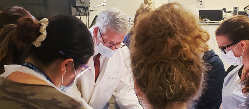 Several Duke University School of Nursing students crowd around a clinical table and listen to a faculty member wearing a white doctor coat. Bright overhead lights shine on subject.